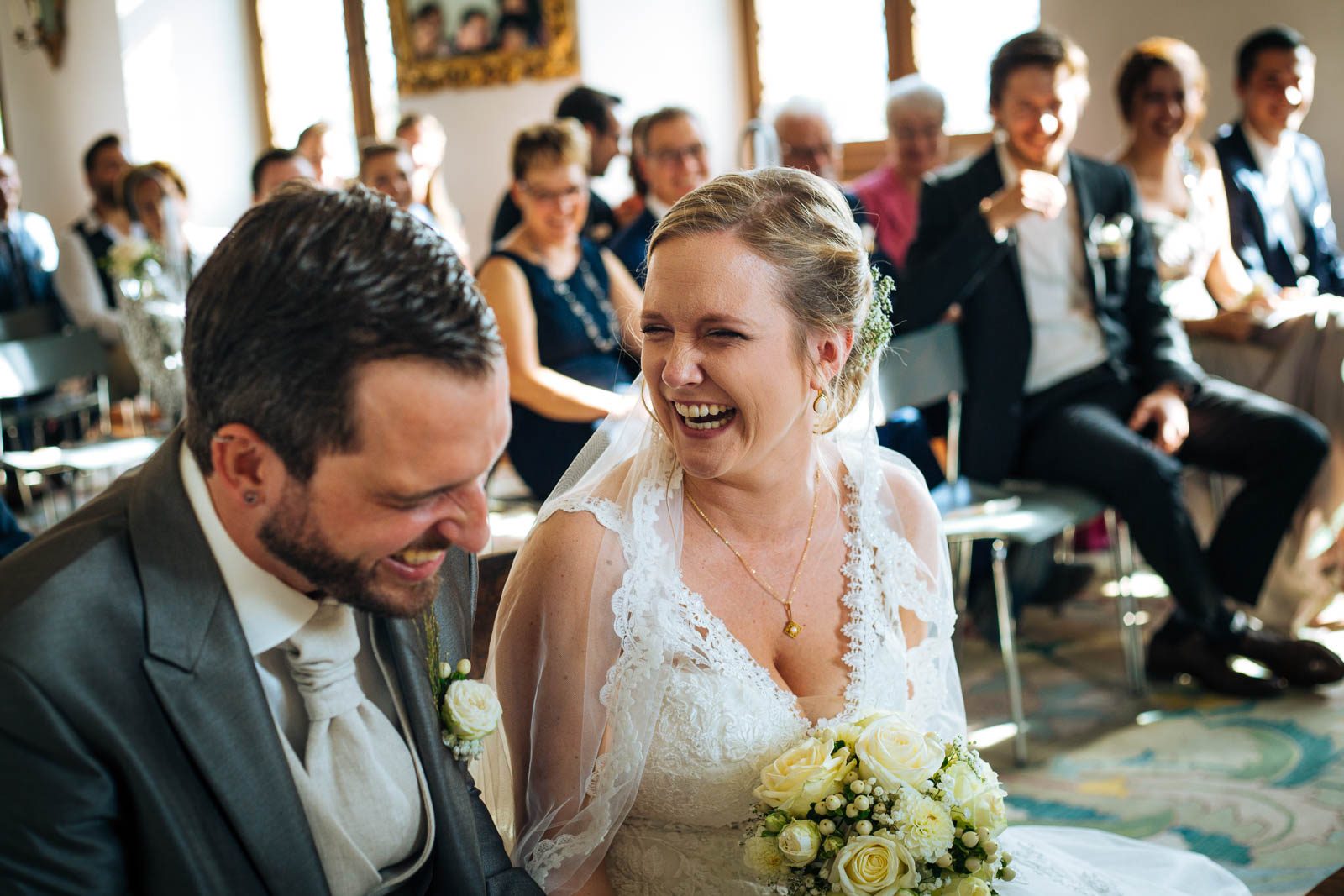 Brautpaar lacht herzlich während der Trauung, Gäste lachen im Hintergrund
